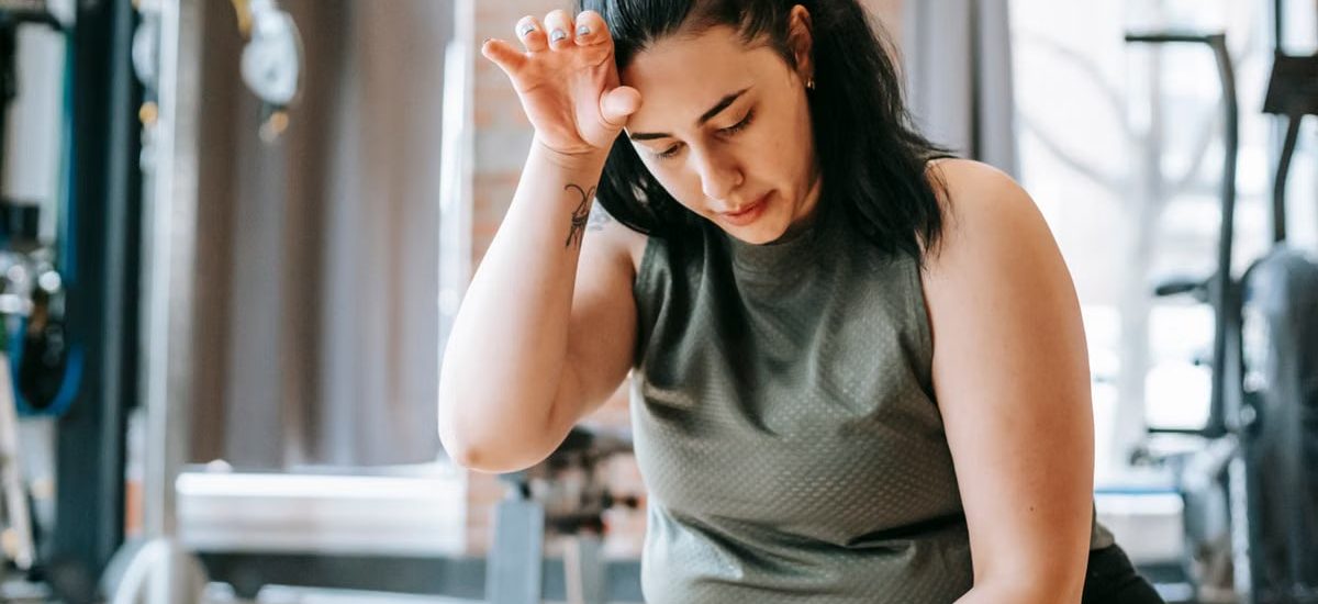 Overweight woman with dark hair in sportswear wiping sweat from face while training in gym in daytime