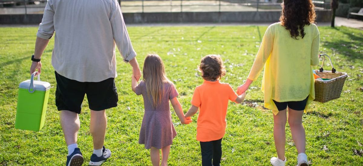 A family of four walking hand in hand through a sunny park carrying picnic supplies.