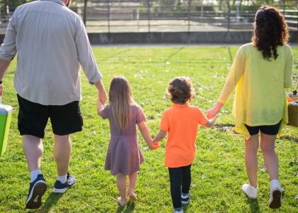 A family of four walking hand in hand through a sunny park carrying picnic supplies.