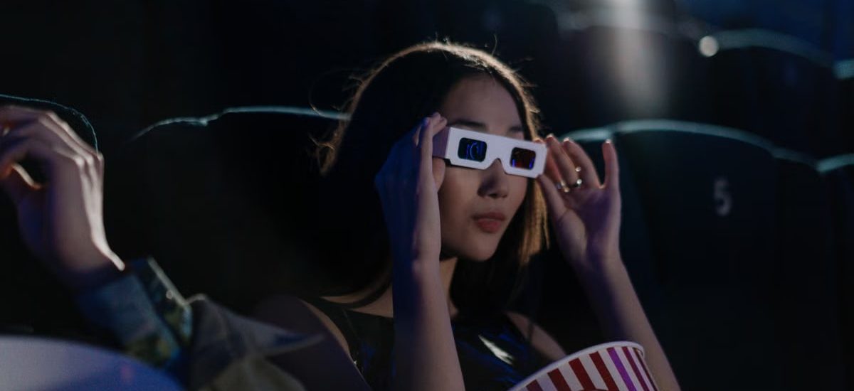 Asian woman wearing 3D glasses, enjoying popcorn in a movie theater.